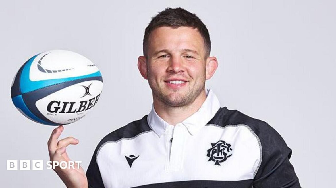 Elliot Dee poses for a camera shoot in a black and white barbarians jersey balancing a rugby ball on the fingers of his right hand