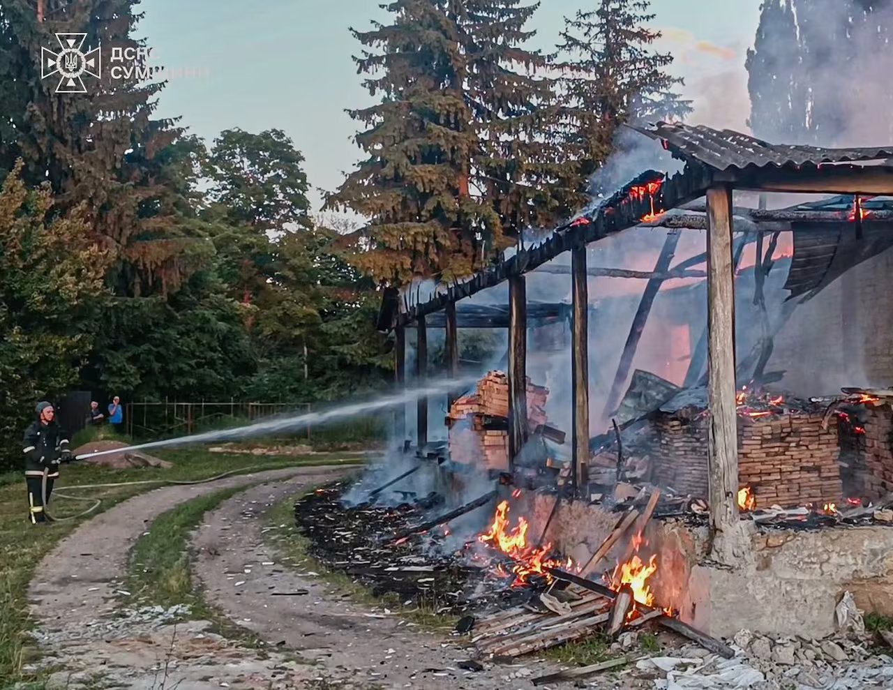 A fireman with a hose is serving a burning structure on a gravel road.