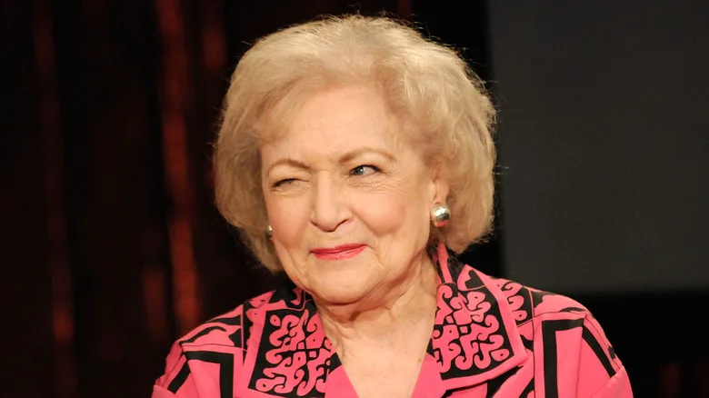 Betty White, wearing a pink and black patterned shirt, winks at someone off-camera.