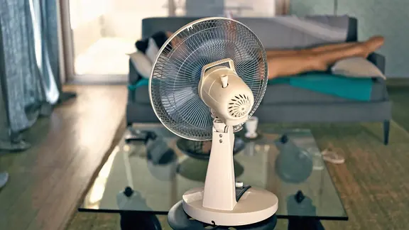 A woman lies on the sofa in front of a fan.