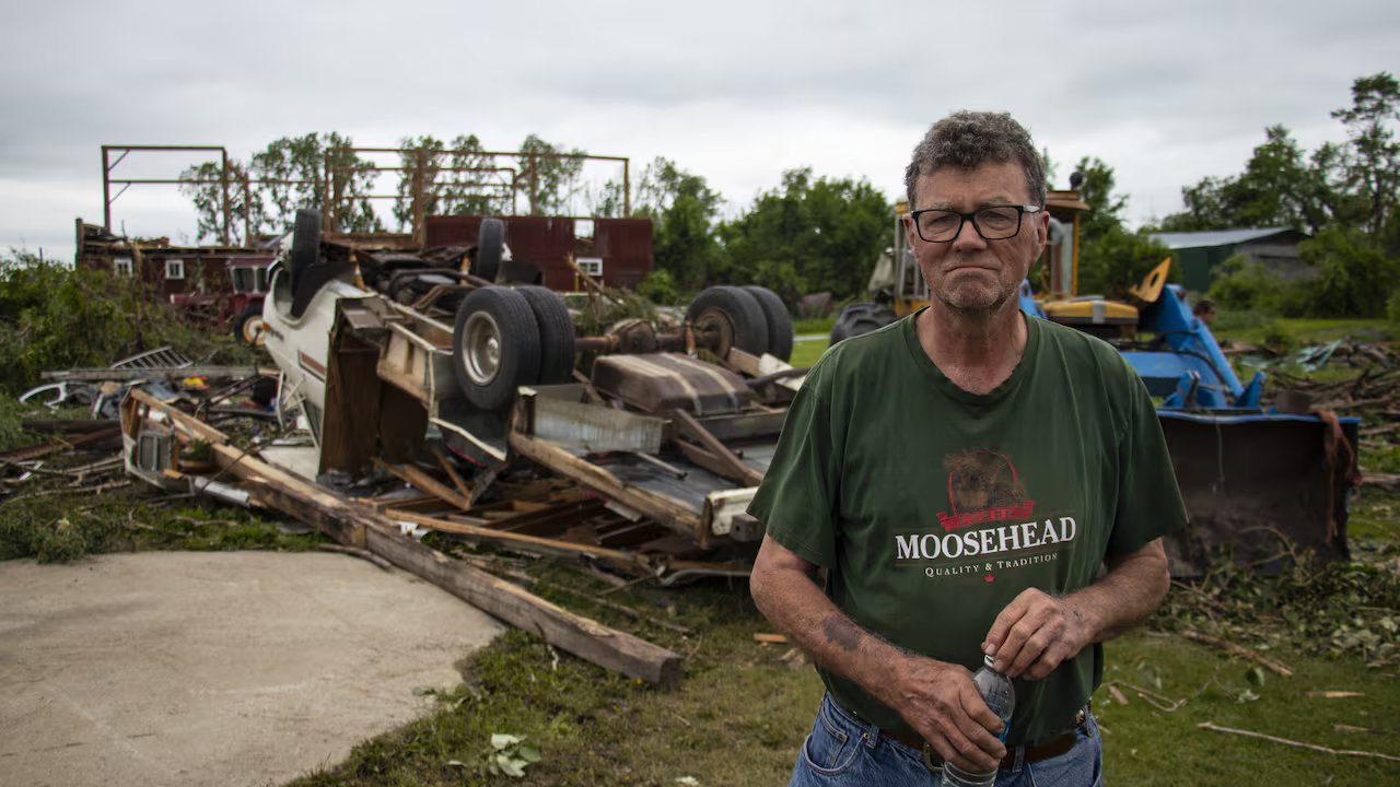 A man stands in front of an overturned car, with debris scattered around.