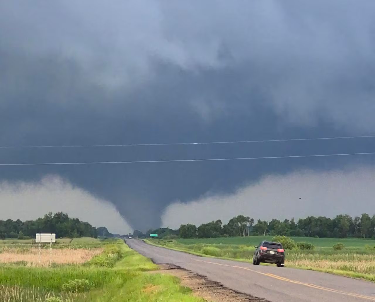 In the distance you can see a deep blue sky and a tornado looking down on a highway with green fields on either side.