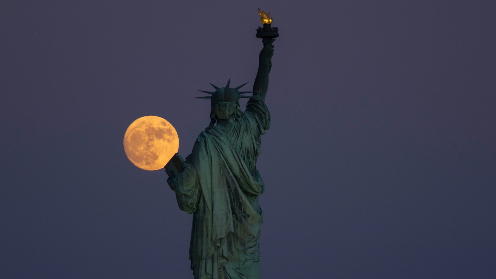June's Strawberry Moon will be a welcome treat for skywatchers around the world. Here are our favorite photos 21 The full moon appears to the left of the Empire State Building, with people sitting outside in the foreground. There are lamp posts decorated with lights and two American flags are visible.