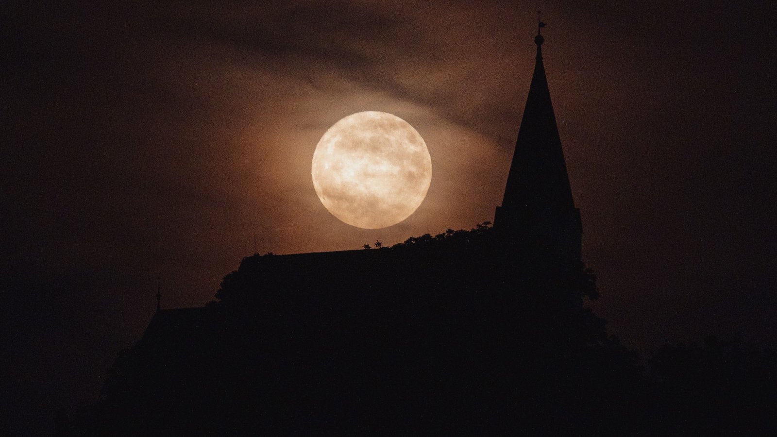 June's Strawberry Moon will be a welcome treat for skywatchers around the world. Here are our favorite photos 28 a fully illuminated moon behind wispy clouds and the silhouette of a church below.