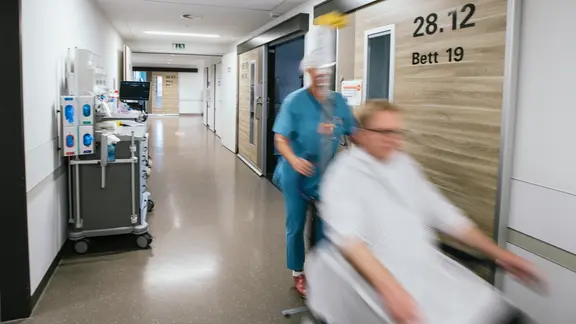 Medical staff pushes a patient down a hospital corridor. | NDR, Julius Matuszczyk Medical staff pushes a patient down a hospital corridor.