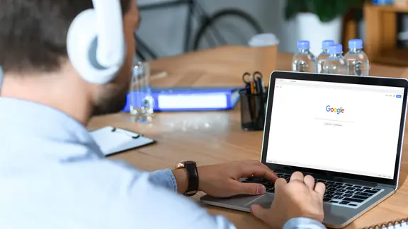 A man sits in front of a laptop with headphones and shows the Google website | Color box A man sits in front of a laptop and shows the Google website using headphones.