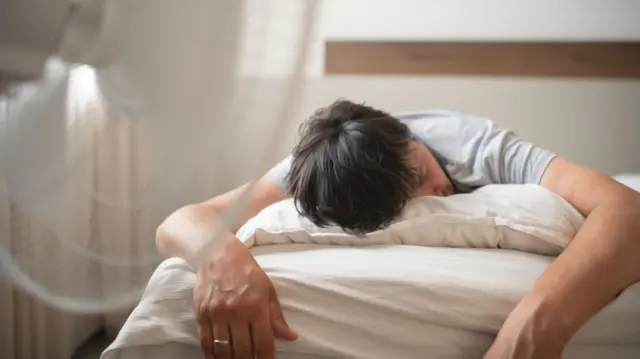 A man hit his head face down on a pillow while sleeping in bed. The fan in the foreground of the picture is directed towards him.