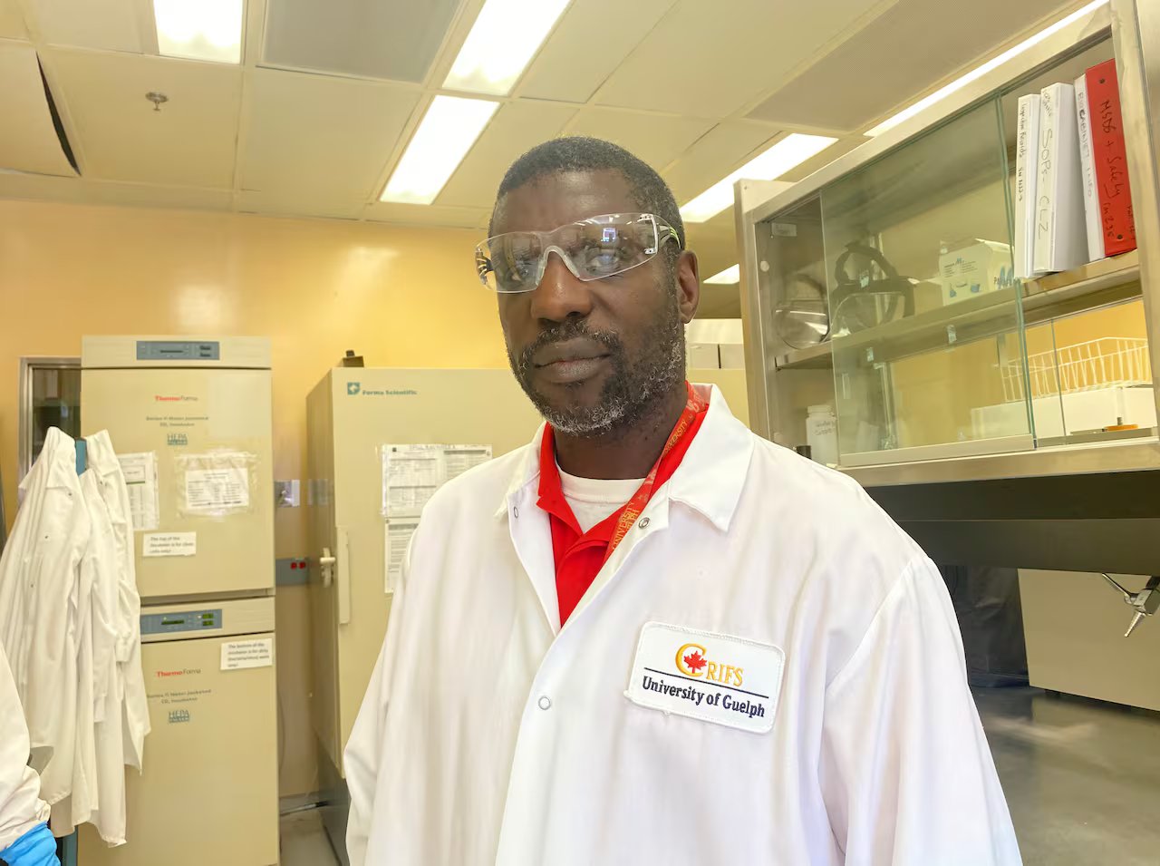 The man stands wearing glasses, a red shirt and a lab coat with an embroidered label on the chest with the words RIFS and the University of Guelph underneath it. 