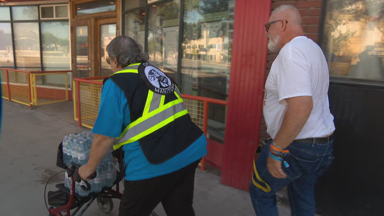 A woman with a walker filled with cases of water bottles walks down a city street with a man in a white T-shirt.