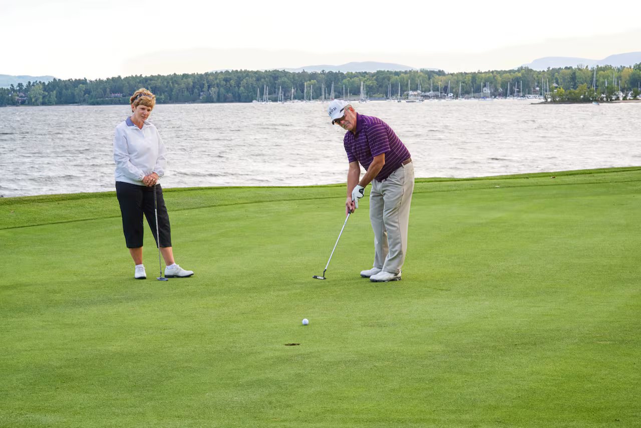 An elderly woman in a black capri and gray sweater stands to the left and watches an elderly man in a red polo shirt and white cap hit a golf ball into the hole. a pond is visible in the background behind the couple
