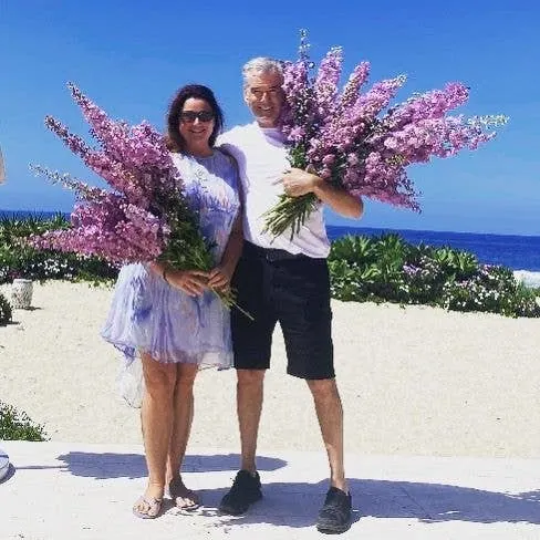 Keeley and Pierce Brosnan carry bouquets of lavender while walking on the beach.
