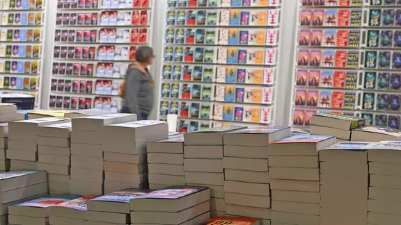 Many paperback books lie on stacks at the Frankfurt Book Fair - in the background a woman stands in front of colorful covers. image Alliance/dpa | Arne Dedert Many paperback books lie on stacks of books at the Frankfurt Book Fair - a woman stands in front of the colorful covers in the background.