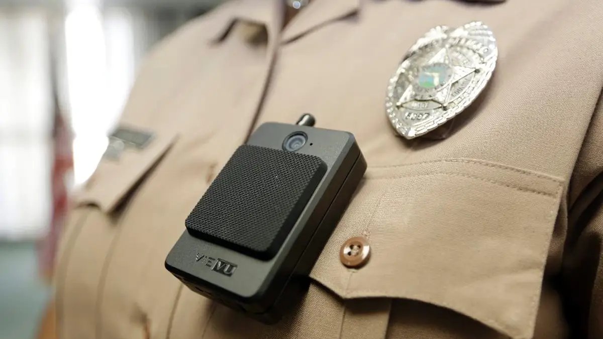 A close-up of a police officer's uniform shows a body camera attached to the front of his shirt under his law enforcement badge.