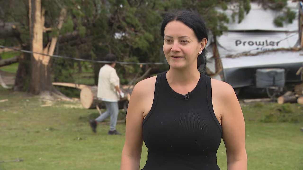 A woman with black hair in a black T-shirt looks at the camera. Behind her are many fallen trees and a man in gloves preparing to clean up.