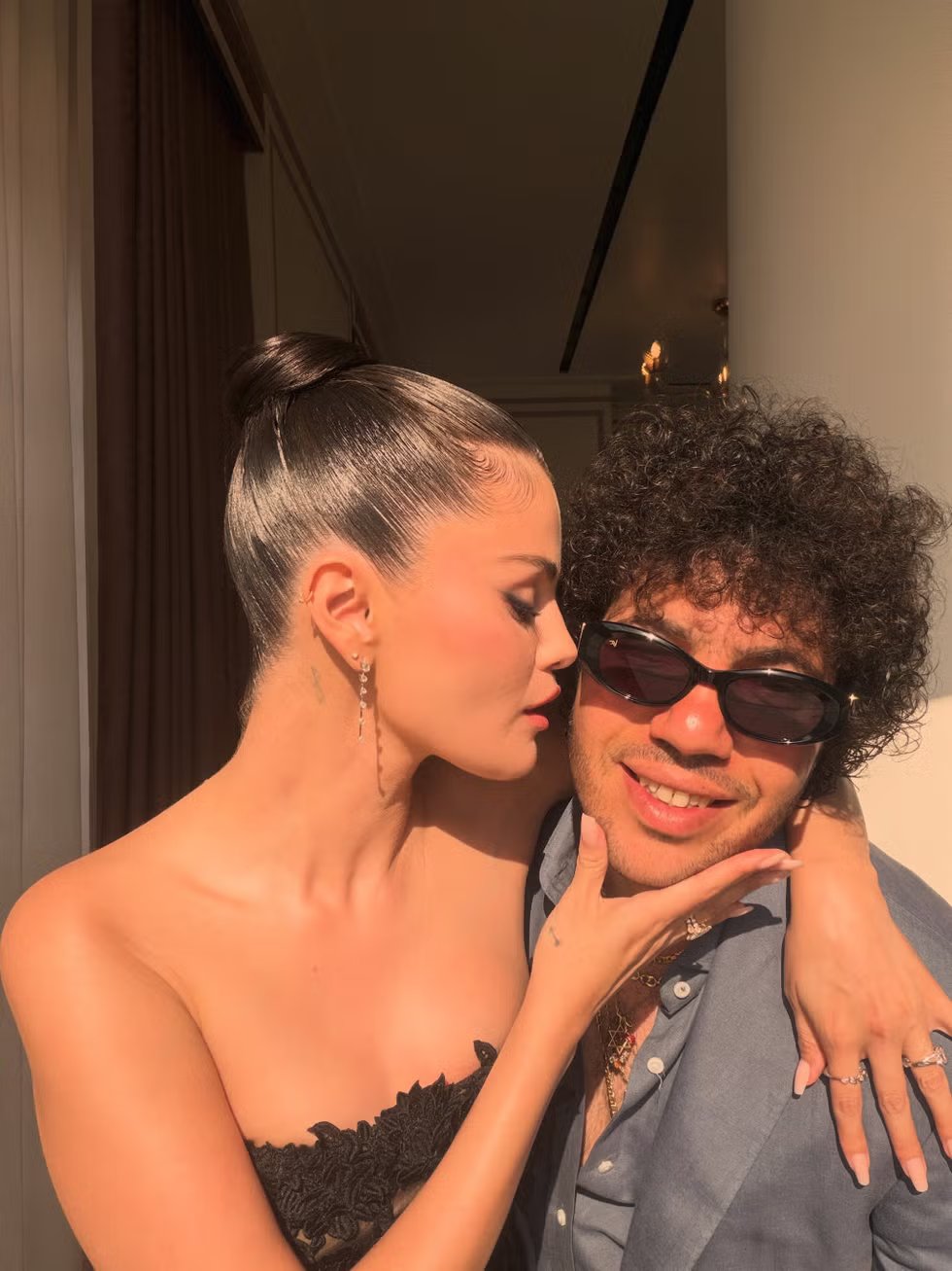 A woman in a black dress leans in to kiss a man in sunglasses, both smiling against a bright background.