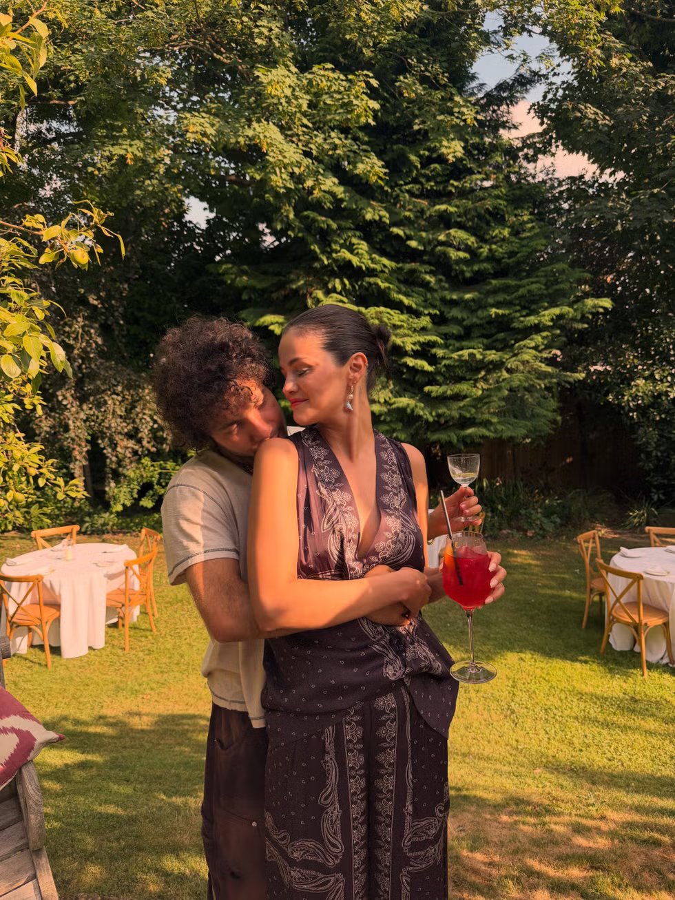 A couple enjoys a get-together in a summer garden, one holding a cocktail and the other hugging.