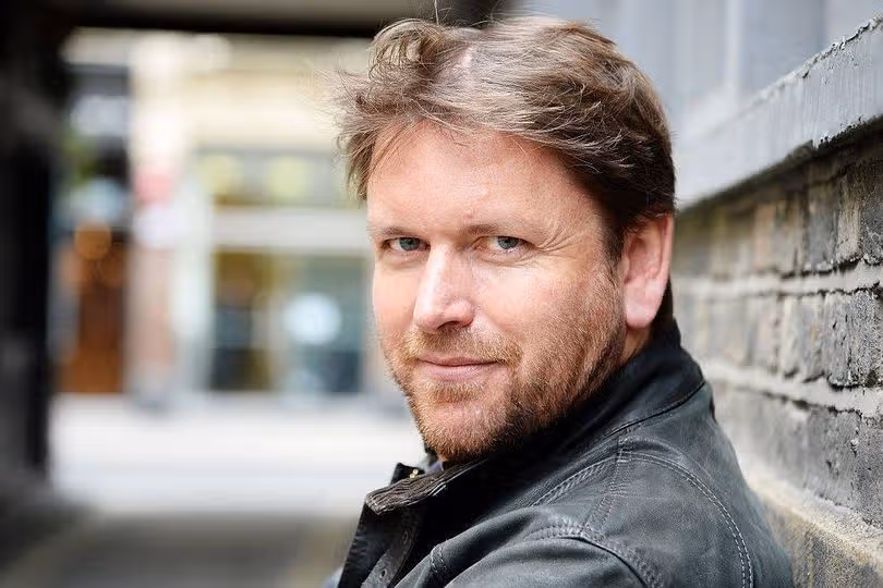 “We visited James Martin's house - Paul Hollywood's hilarious mockery made us shudder.” 14 A man with a beard in a black jacket stands next to a brick wall, smiles and poses for a photograph. In the background is a blurry view of a street with several buildings and institutions.