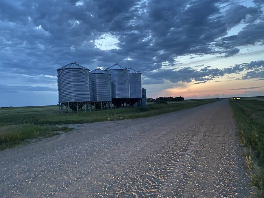 Grain bins next to a gravel road, the sun behind the clouds and setting in the background.