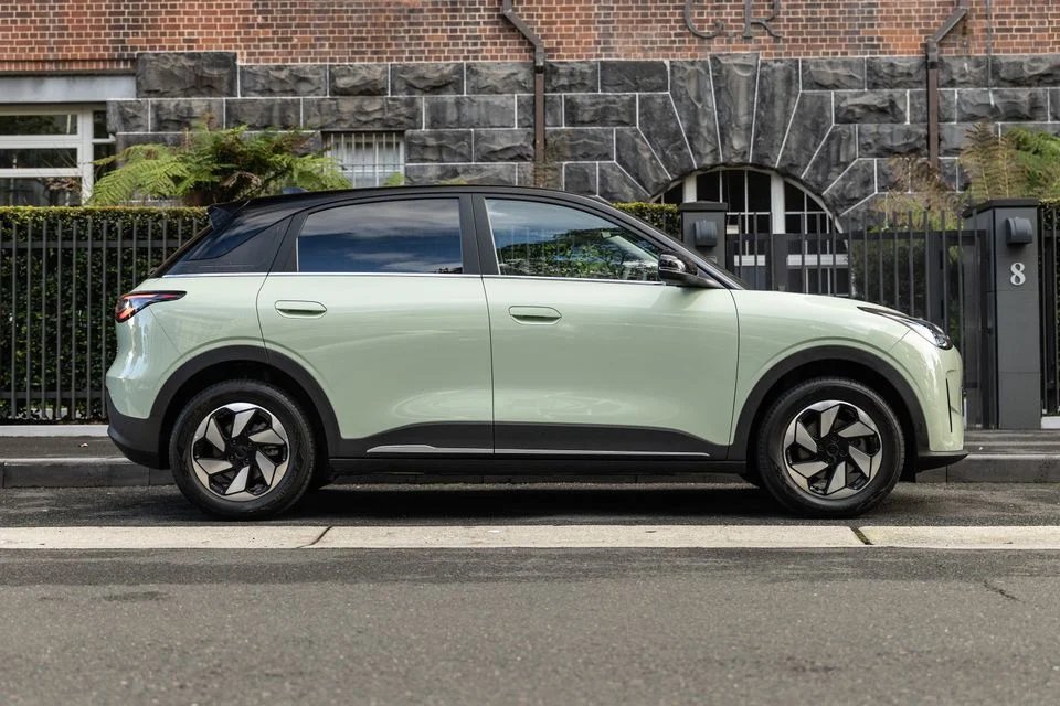 Light green Geely EX2 small SUV, side view, parked on the street in front of a brick building.