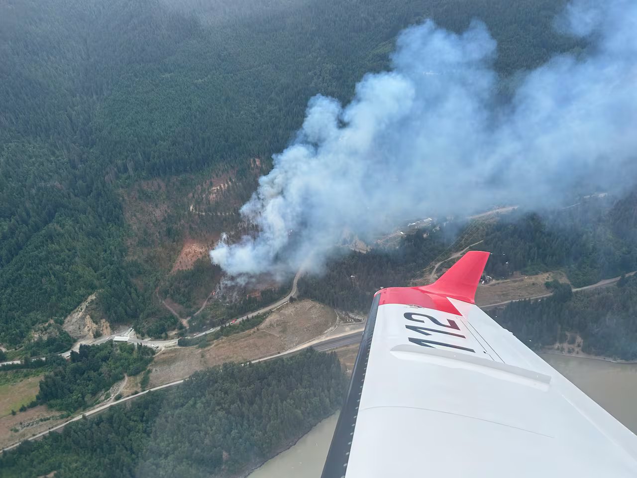 Aerial view from an airplane: smoke rises from a forest fire burning on a forested mountainside near roads and a river.