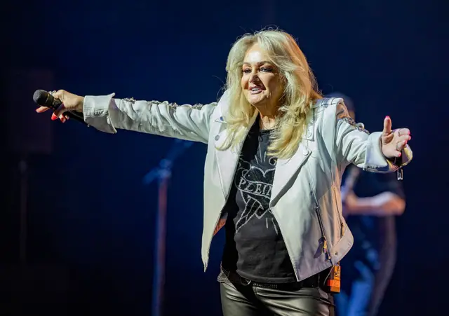 Bonnie Tyler performs on stage with a smile on her face and her arms wide open. She holds a microphone in her right hand.