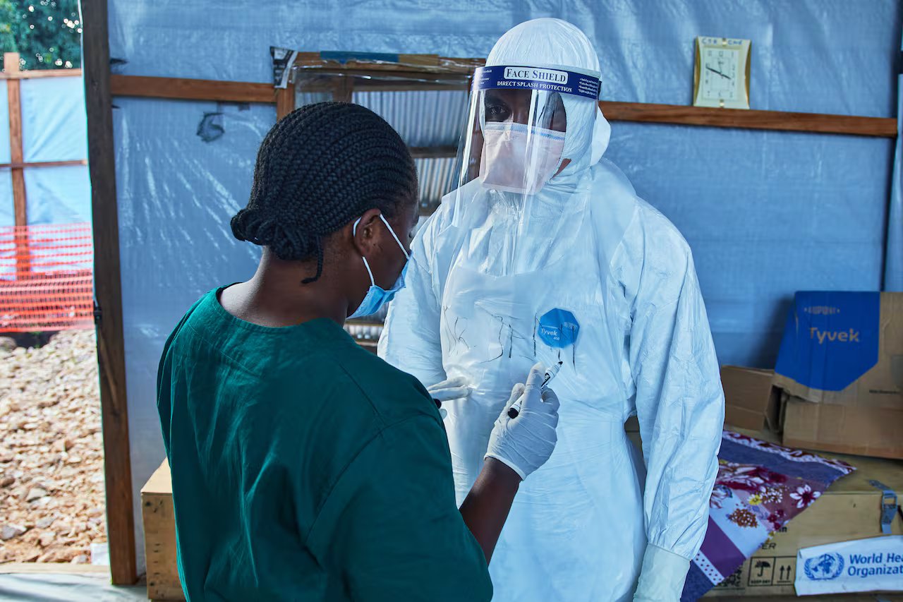 Two medical workers, one of whom is wearing a protective suit, including a protective mask, are in a makeshift tent.