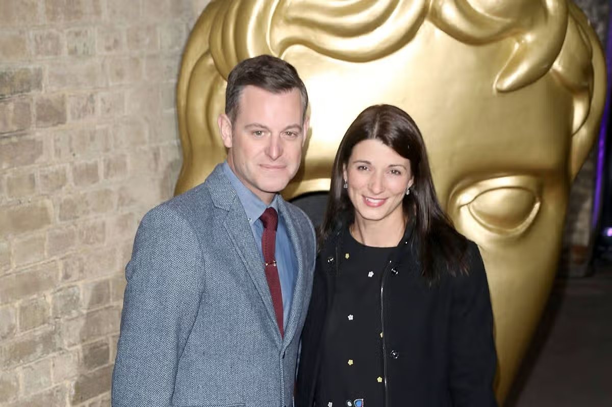 A man and woman, both formally dressed, stand side by side in front of a large golden head sculpture. The setting suggests a formal event or awards ceremony.