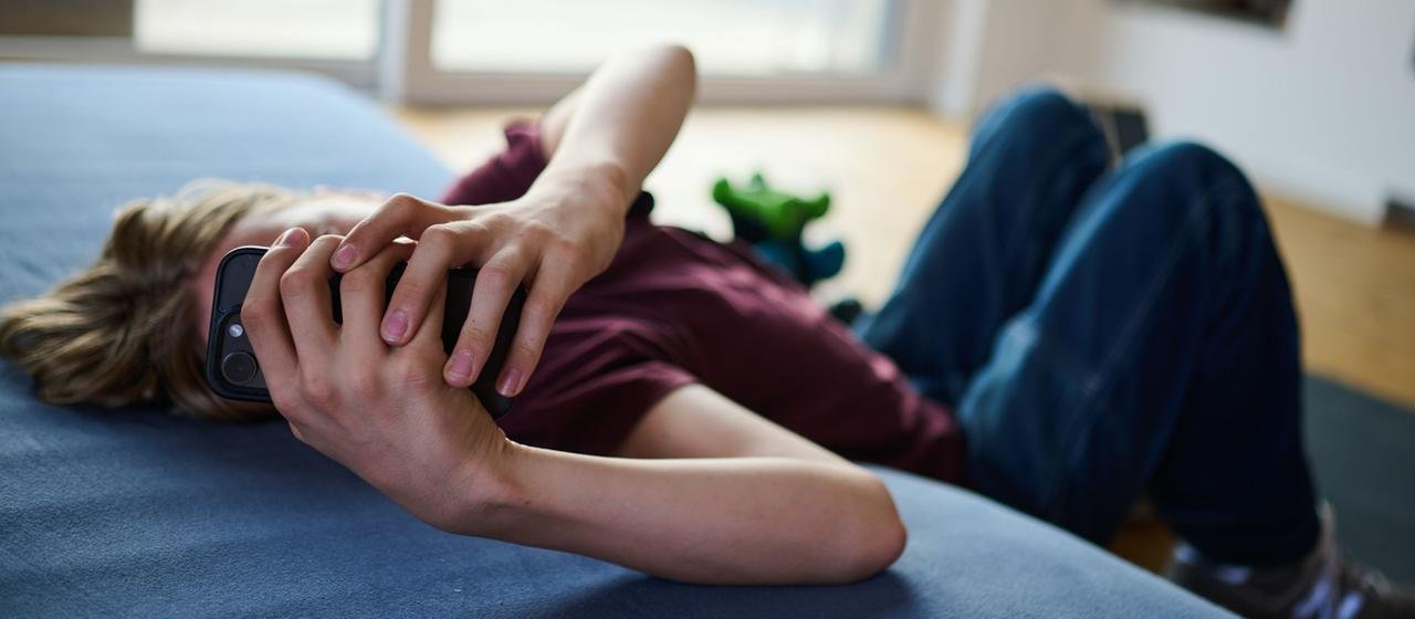 A teenager looks at a mobile phone while lying on the floor and mattress. | Annette Riedl/dpa A teenager looks at a mobile phone while lying on the floor and mattress.