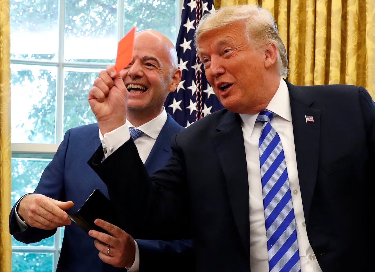 Two men in suits smile while standing together near the window.