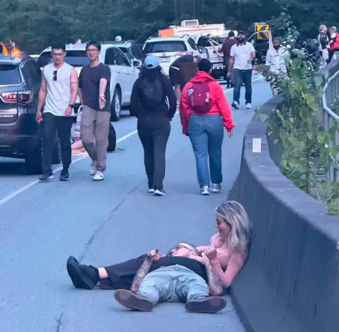 A highway with a line of stopped cars. People walk on the shoulder. In the foreground, a woman sits on the ground with a man's head in her lap.