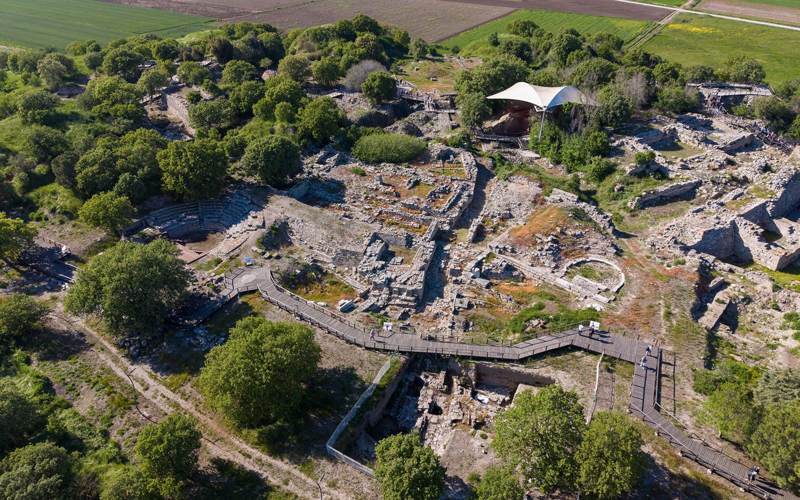 Aerial view of an archaeological site surrounded by green trees and fields, wooden walkway along the facade