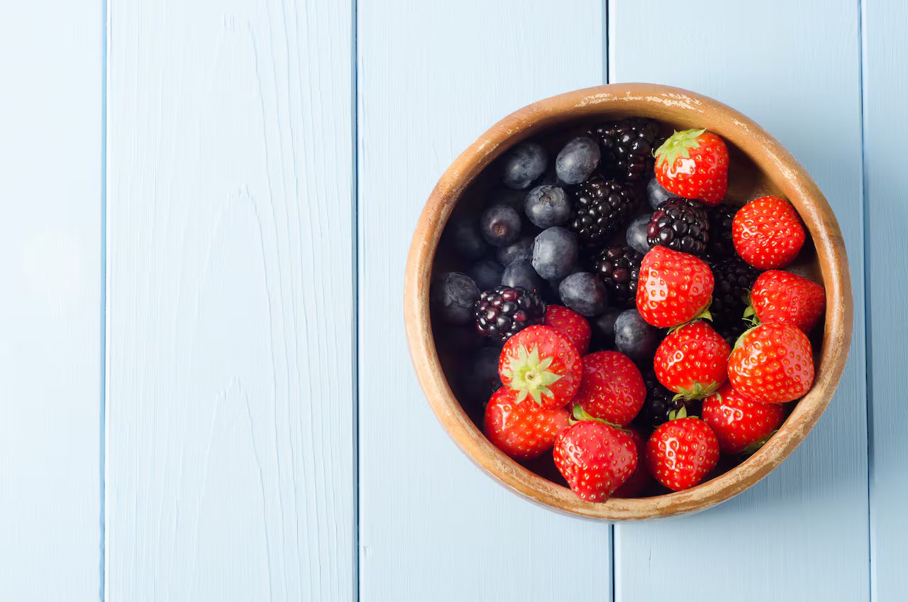 Wooden bowl with strawberries, blueberries and blackberries on a pale blue wooden table.
