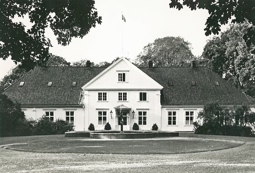 Historic manor house with symmetrical windows and a central entrance surrounded by trees.