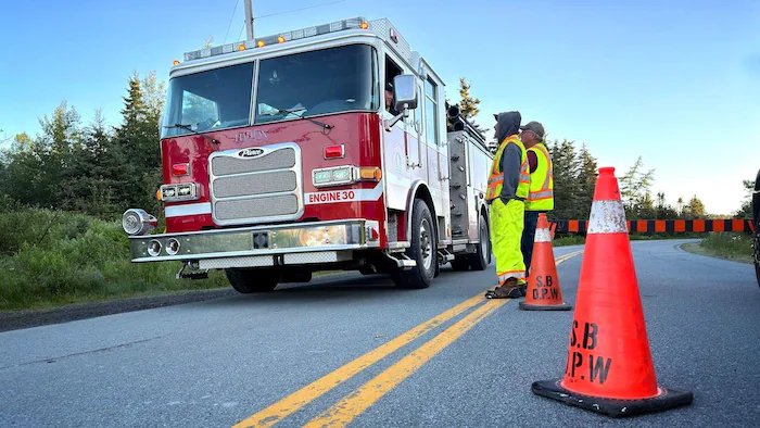 Two men in reflective yellow uniforms stand on a rural road and watch a fire truck slowly approach.