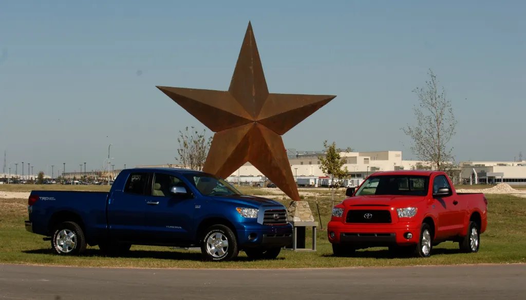 Two Toyota Tundra pickups, one blue and one red, are parked in front of a large metal sculpture of the star, with an assembly plant in the background.