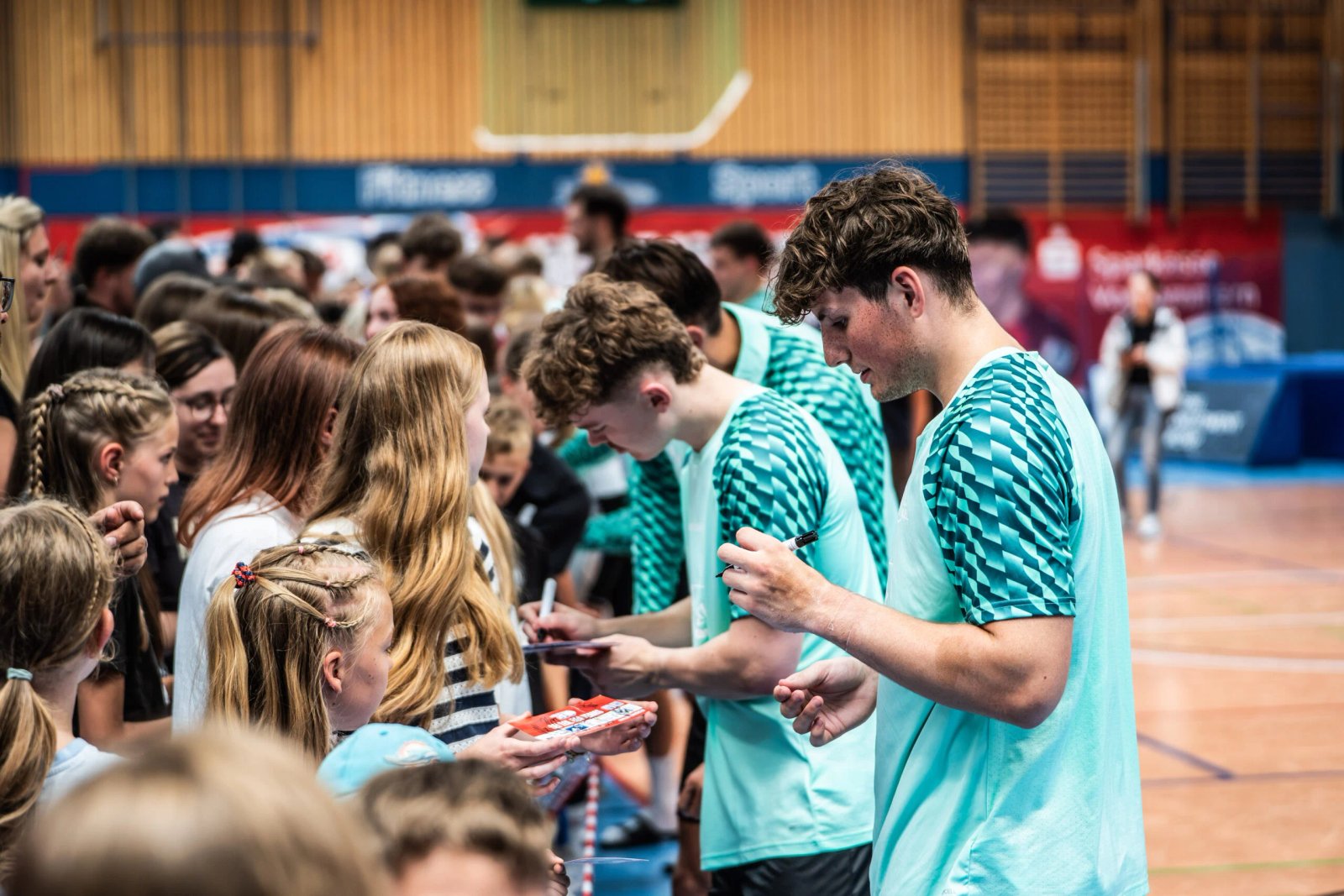 Several athletes in blue and green T-shirts sign autographs to a crowd of young fans in the gym. Activity is buzzing and fans eagerly hand out items to sign - the lively atmosphere is reminiscent of the moment when new coach Børge Lund was introduced at THW Kiel.