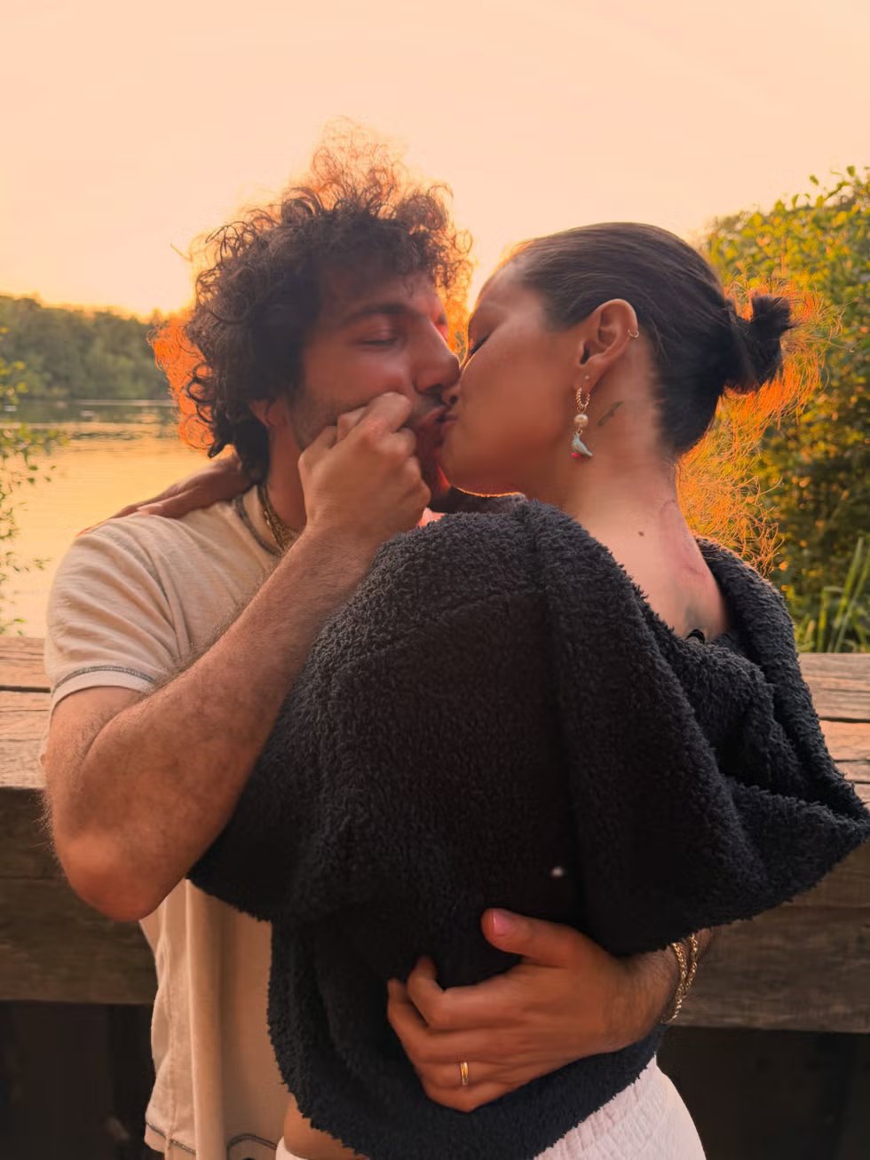 A couple kisses romantically at sunset on the shore of a lake, surrounded by nature.