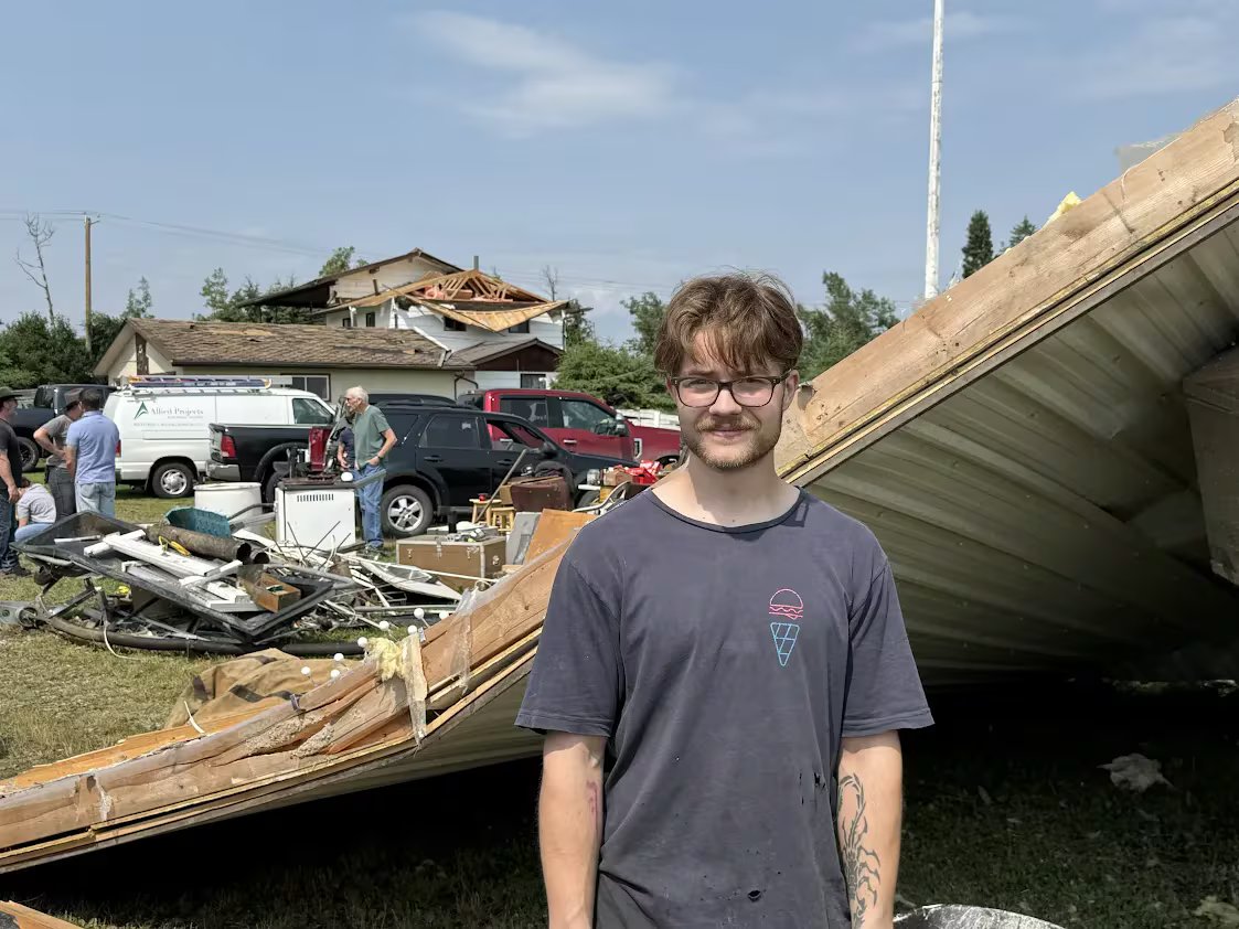 A man with brown hair and a bard wearing glasses and a T-shirt stands in front of a house damaged by a tornado.