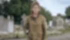 A woman in an olive jacket stands in a cemetery with tombstones and looks pensive under a cloudy sky.