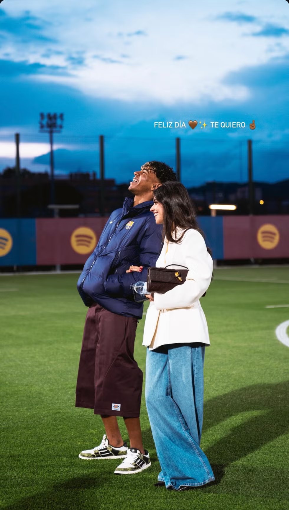 Two people smile and stand together on a football field with a message hanging above them commemorating their special day.